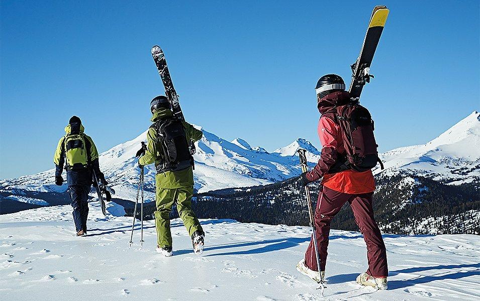 Three skiers walk up the mountain in a single file line on a gorgeous sunny day. 