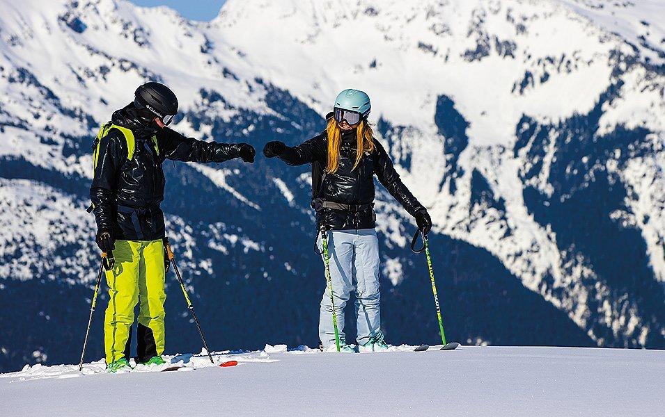 Two skiers reach out and bump fists as they smile on top of a ski run. 
