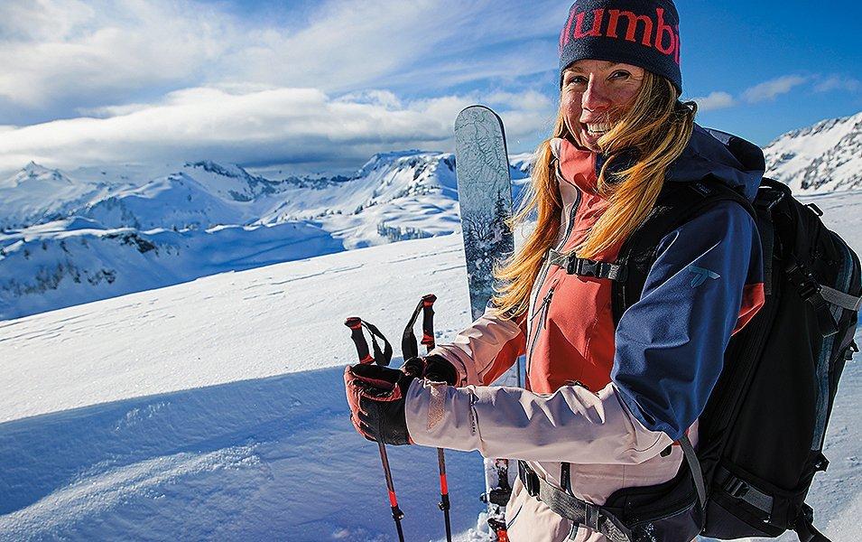 A skier stands in front of a giant cornice of untouched powder as she looks at the camera and smiles.