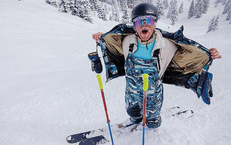 A skier with snow caked in his mustache removes his ski coat as he stands on a powdery ski run. 
