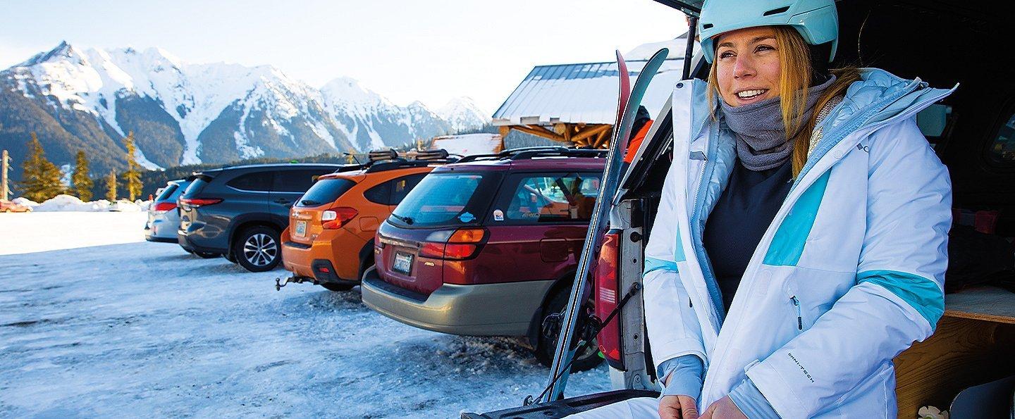 A woman stands in the parking lot of a ski resort zipping up her jacket.  
