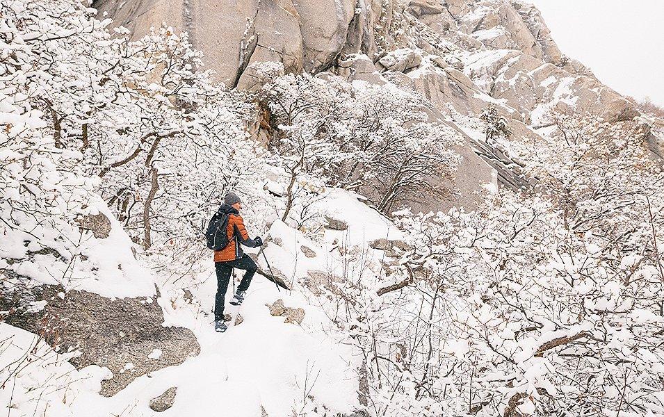 A bundled-up man in an orange puffy jacket and black snow pants stands with trekking poles on a snowy cliff watching the scenery.