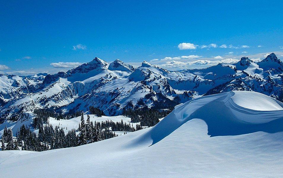 A snow-covered valley full of deep powder sits in the foreground of Mount Rainier National Park while gorgeous mountains sit in the background under a bright blue sky filled with fluffy white clouds.