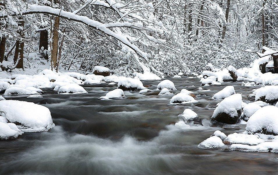 Snow-covered boulders line a flowing river as it rushes through a winter landscape in Great Smoky Mountains National Park.