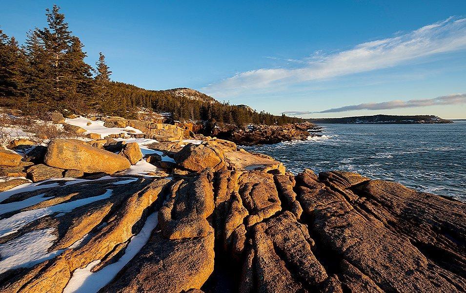 Golden rocks covered with pockets of snow sit next to the deep blue ocean as the morning light hits them in Acadia National Park.