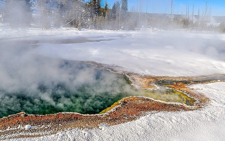 A misty geyser with green and yellow water that’s surrounded by snow emits steam in Yellowstone National Park.