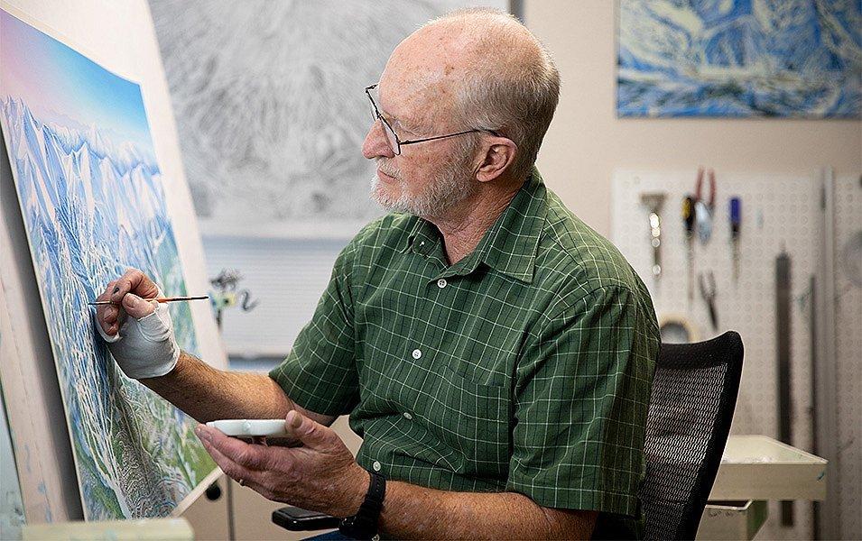 James Niehues sits at his easel holding a paintbrush and smiling for the camera.  