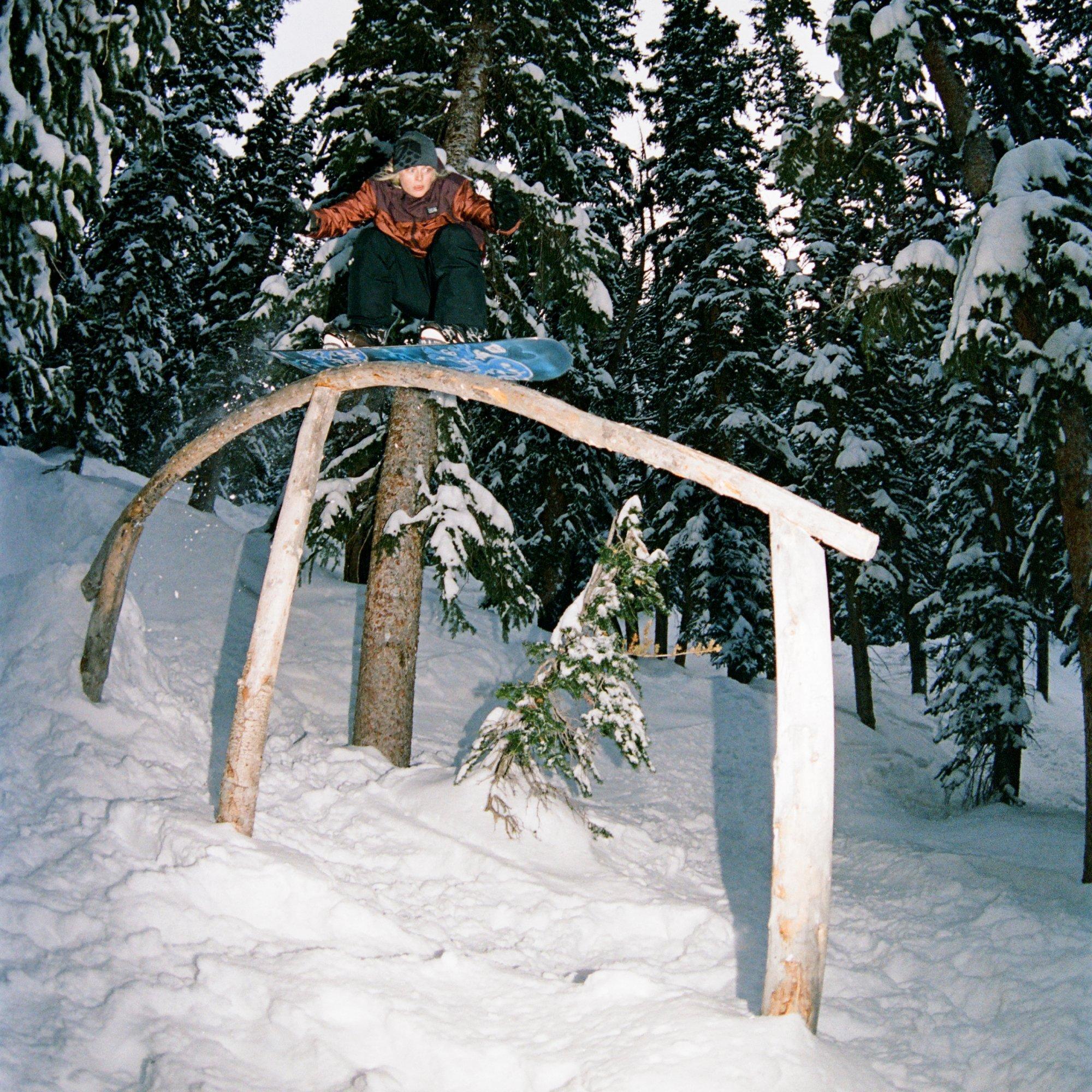Stine Tønnessen glides over a tree branch ramp on her snowboard through a snowy forest.