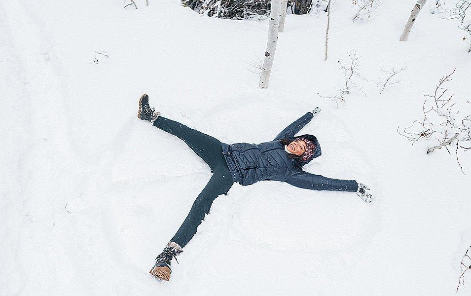 A woman wearing a puffer coat smiles as she makes snow angels on the ground in a winter setting. 