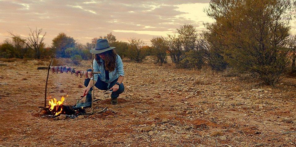 A women cooking bacon over an open fire. 