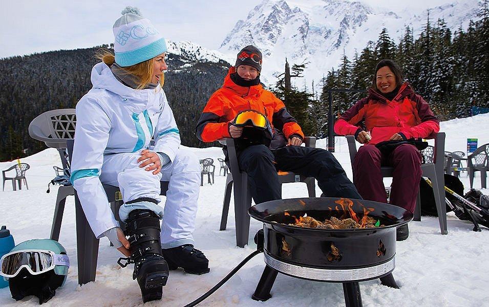 A group of three skiers sit around a firepit on a ski slope with a mountain and some trees in the background. One woman is wearing a white ski jacket with white pants; a second woman is wearing a red jacket and burgundy pants; and a man in the middle is wearing a red jacket and black pants.