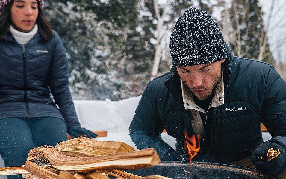 A man crouches down next to a campfire in a scenic winter setting as a woman looks on in the background.  
 