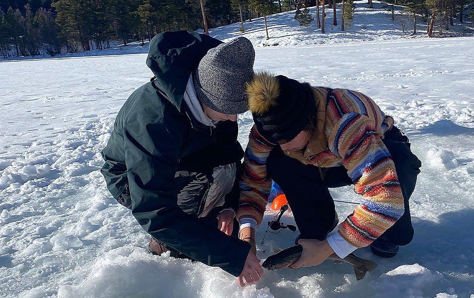 A kid in a multi-colored striped sweater and black hat crouches in the snow over an ice hole next to a woman wearing a black jacket and gray hat. 
