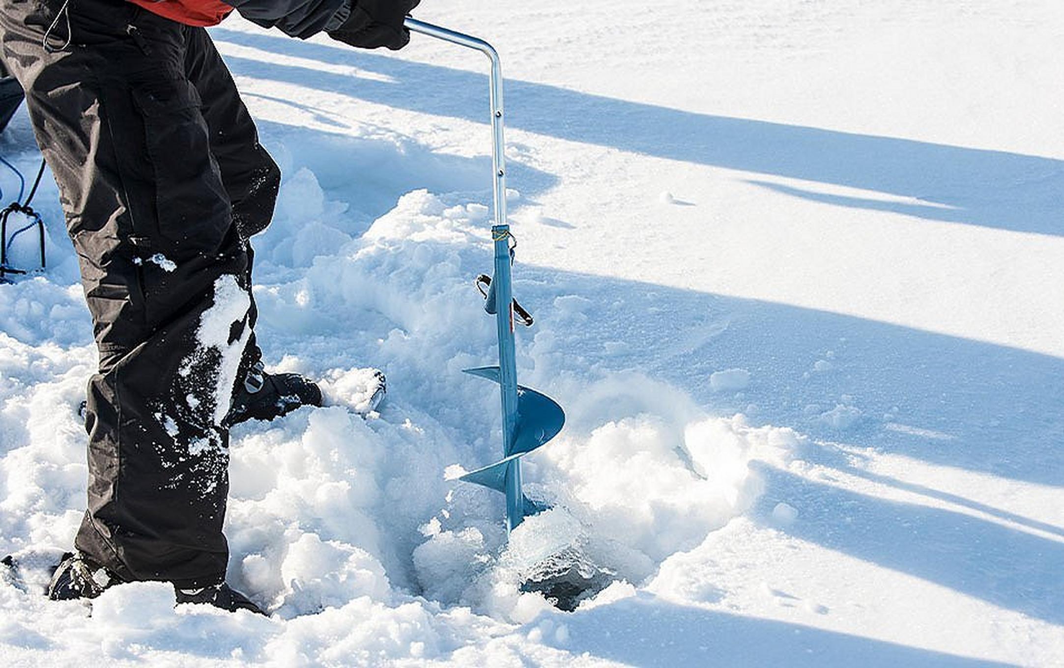 A close-up photo of a person pictured from the waist down wearing black snow pants using an auger to drill through the ice.
