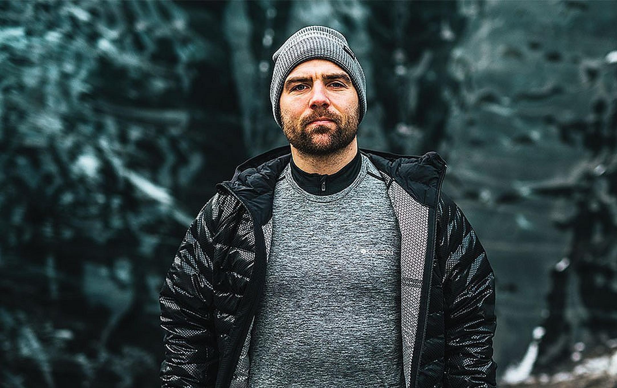 A man wearing an unzipped black Columbia Sportswear jacket with a gray baselayer shirt underneath it stares at the camera with a serious face.