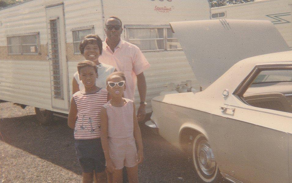 An old snapshot shows Benjamin and Frances Graham sitting on a picnic table outside a travel trailer with a group of people they met on the road. 