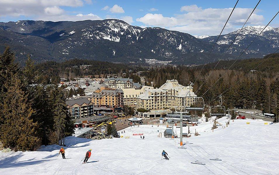 A wide shot of skiers heading down the slopes on a sunny day at Whistler Blackcomb with a ski lodge in the background. 
