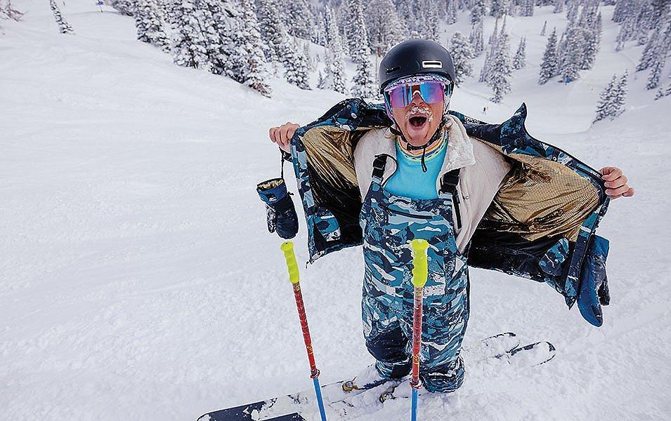 A skier wearing blue camo Columbia Sportswear bibs opens his jacket in a snowy powder field.  