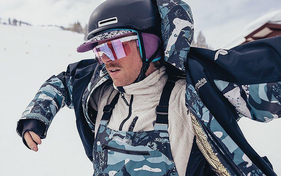 A skier wearing blue camo Columbia Sportswear bibs takes off his jacket in the middle of a ski slope. 