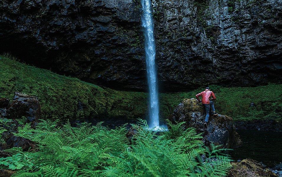 A man wearing a red jacket climbs up a rock in the distance next to a waterfall surrounded by lush green moss and ferns.