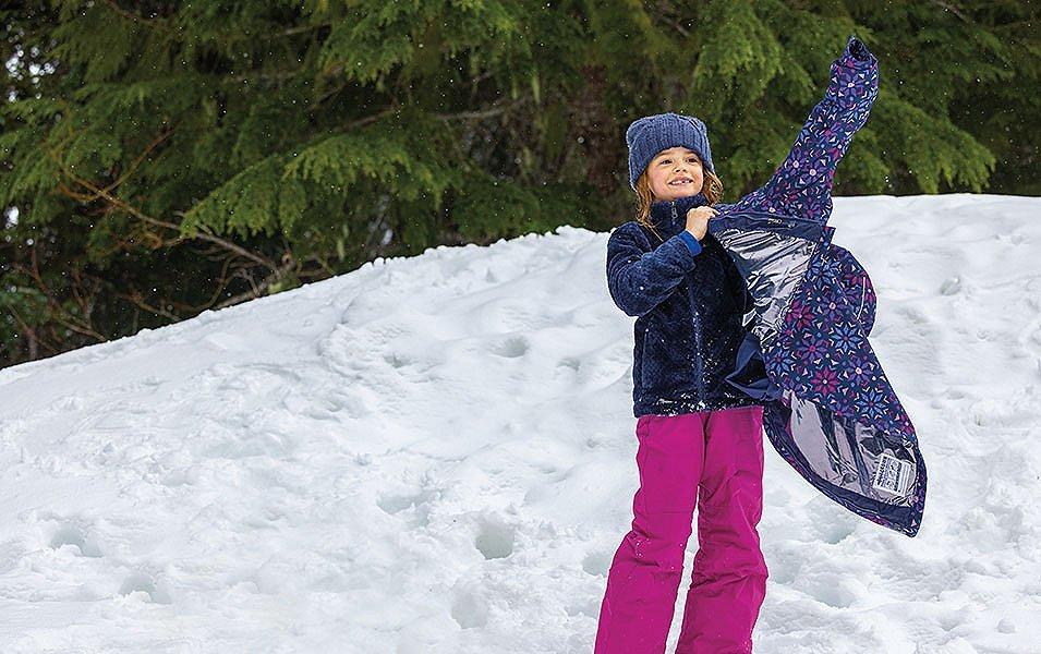 A young skier standing in the snow takes off a purple Columbia Sportswear jacket as she smiles off camera on the mountain.