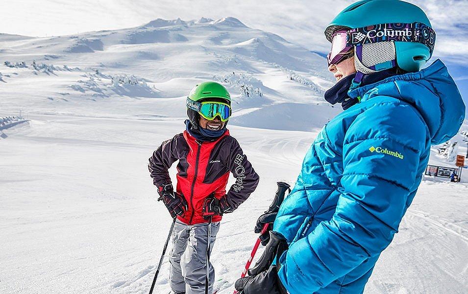 A skier wearing a blue Columbia Sportswear jacket smiles as he talks to a youth standing next to him on a sunny day on a snow-covered mountain. 