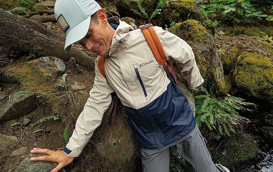 A man hikes alone next to mossy rocks in a lush green setting.