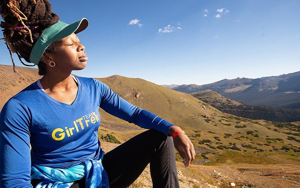 An African American woman wearing a blue GirlTrek jacket basks in the sun in the middle of a beautiful landscape. 