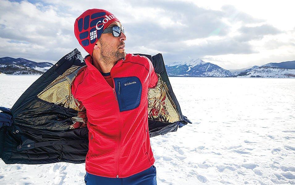A man stands in a scenic snowy setting taking off a Columbia Sportswear jacket with a layer of red fleece underneath. 
