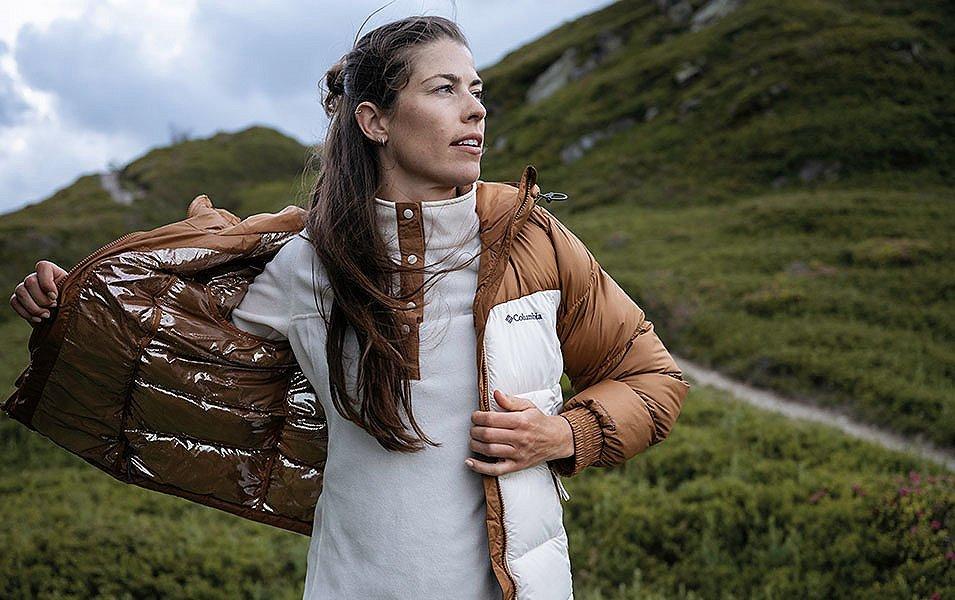 A hiker stands in front of a lush mountain trail taking off a brown Columbia Sportswear puffer coat. Underneath, she is wearing a white fleece midlayer.