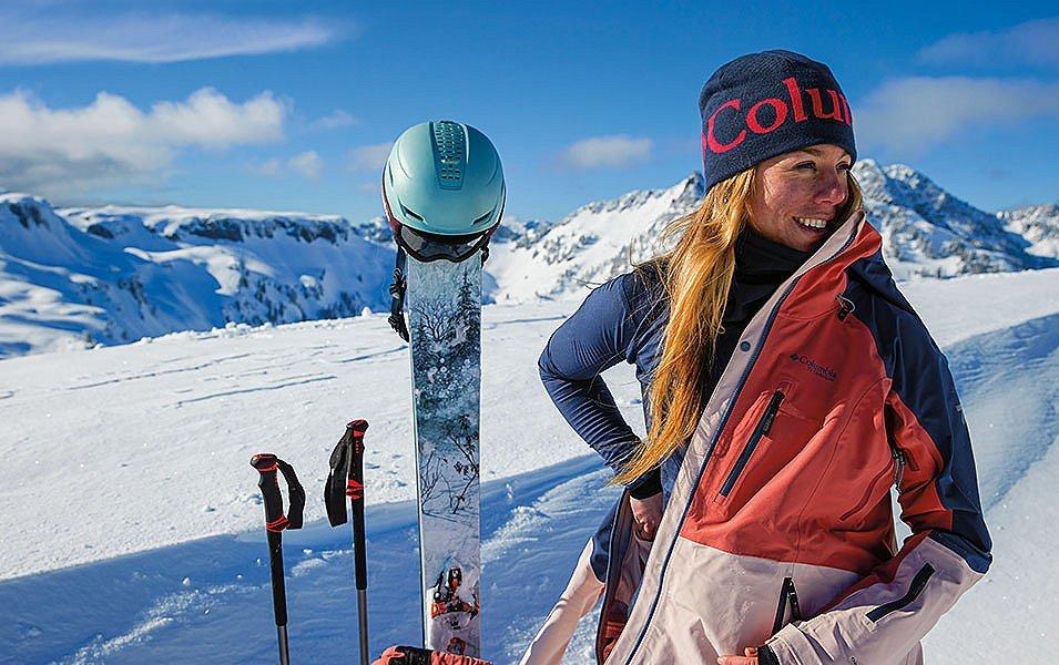 A skier stands on a snow-covered mountain taking off her ski jacket as her skis, poles, helmet, and backpack stick out of the snow next to her. Underneath her ski coat she is wearing a warm Columbia Sportswear baselayer. 