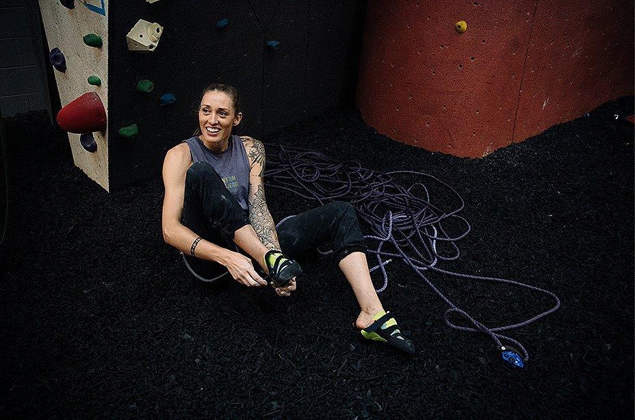 Alex Johnson is sitting on the ground of a climbing gym, putting on her climbing shoes, smiling and looking up, presumably at someone climbing out of sight.
