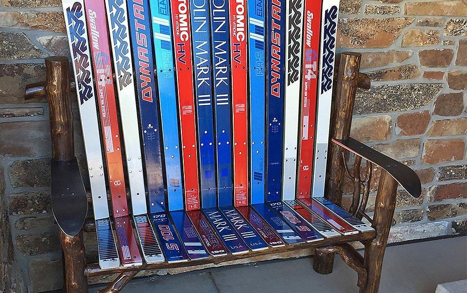 A patio bench made from colorful skis sits outside in a concrete area in front of a brick wall. 