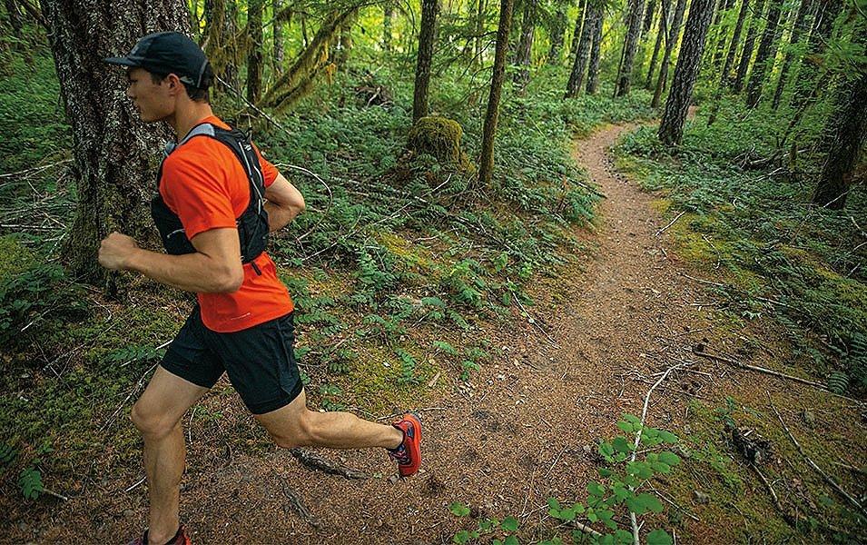A man in a red jacket and a black vest runs on a trail in a scenic forest area. 