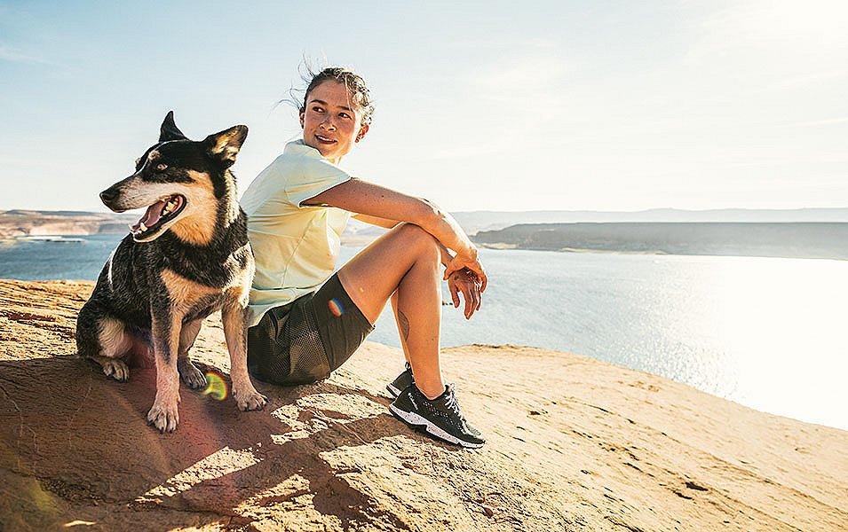 A woman sits on a rocky overlook above a sunny blue body of water with a smiling black dog at her side.