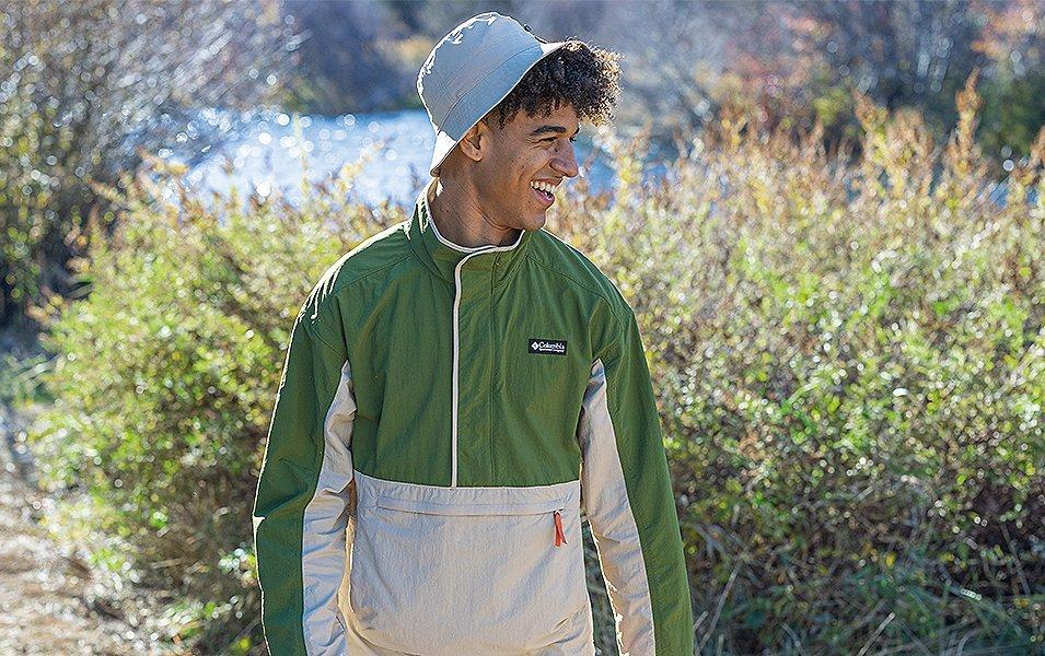  A young man stands in front of a river wearing a beige bucket hat and smiling.