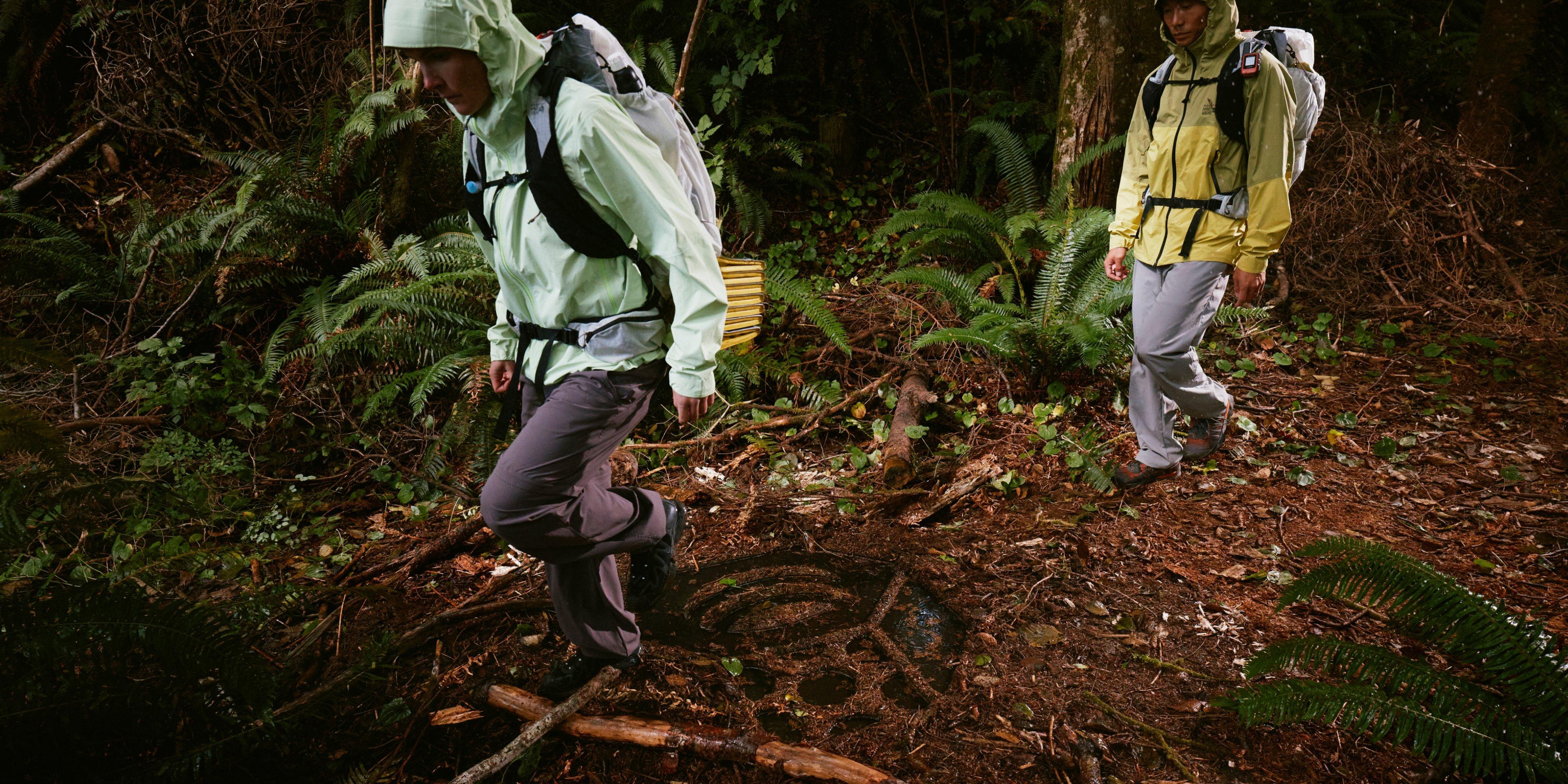 A man and woman in Cloud Cipher™ Hooded Jackets traverse over a Mountain Hardwear nut logo full of water imprinted on the forest floor.