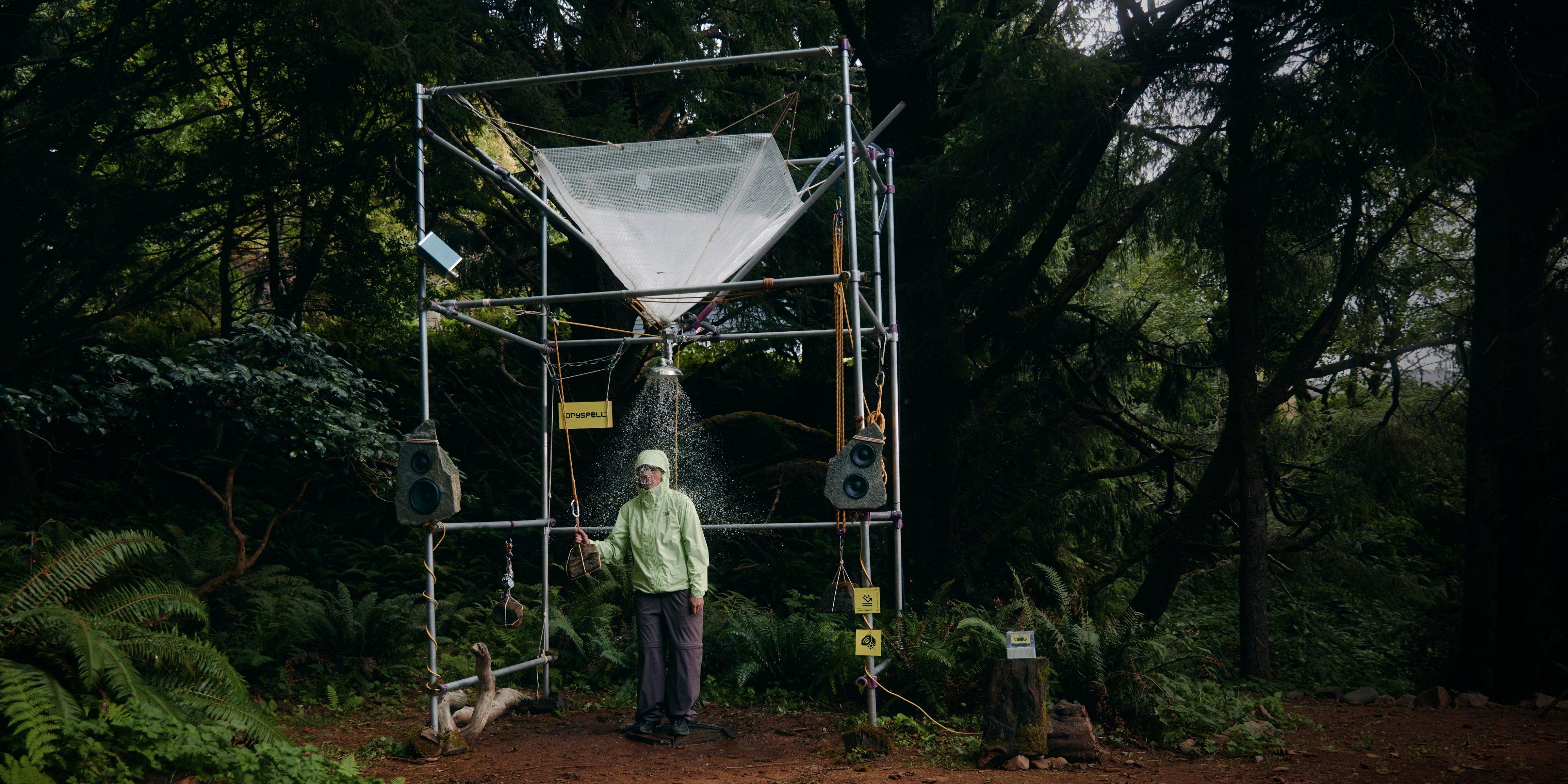 A woman in a zipped Cloud Cipher™ Hooded Rain Jacket stands in a forest clearing under a scaffolding holding a full tarp. She pulls a rope, unleashing a shower of water on her head.