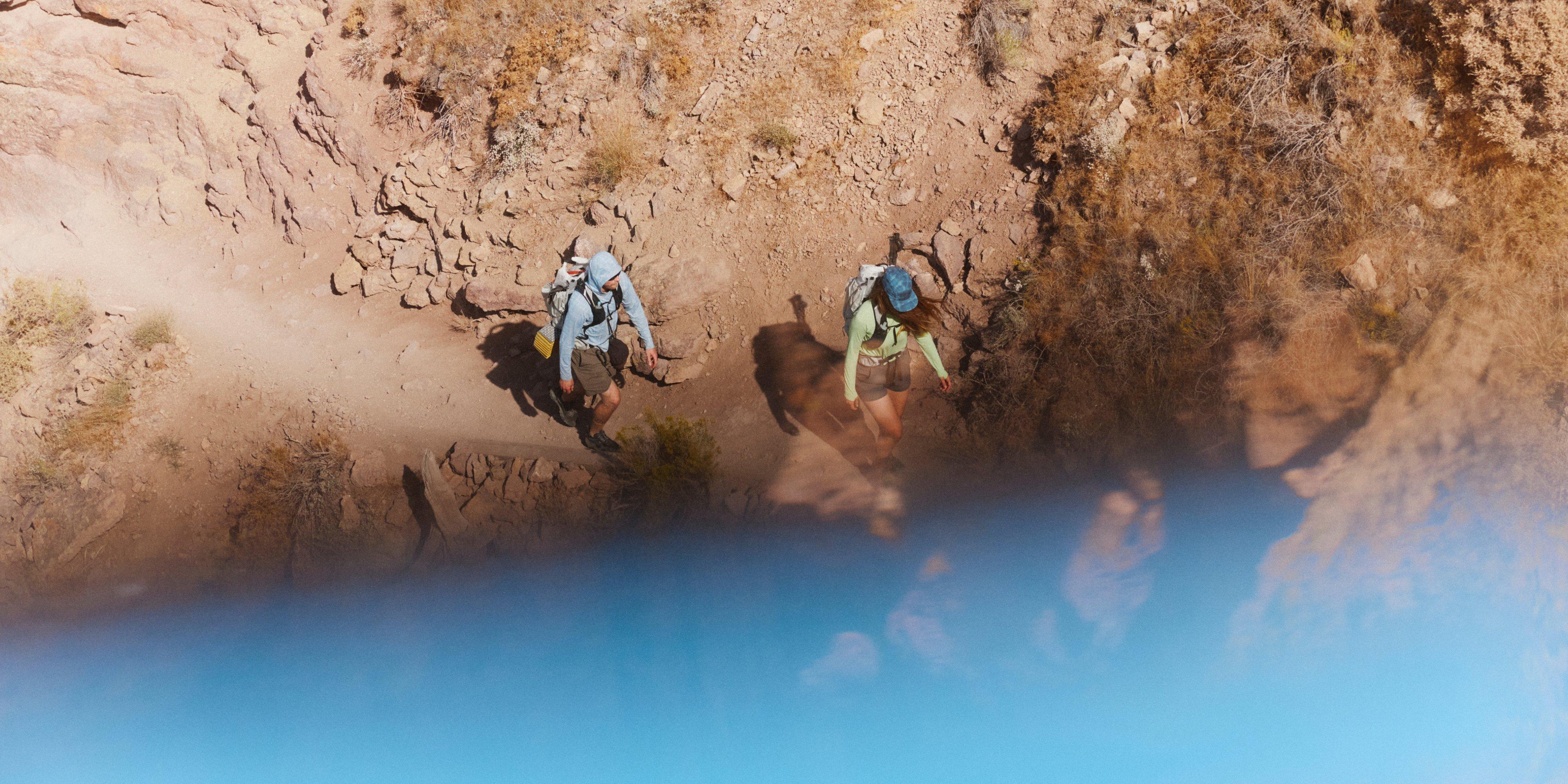 An aerial view of a man and woman in a Crater Lake™ Hoody and Long Sleeve, traversing a the barren dirt and rock path with no shade.
