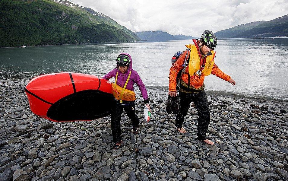 Two people walking on a stony beach carrying kayaking equipment. 