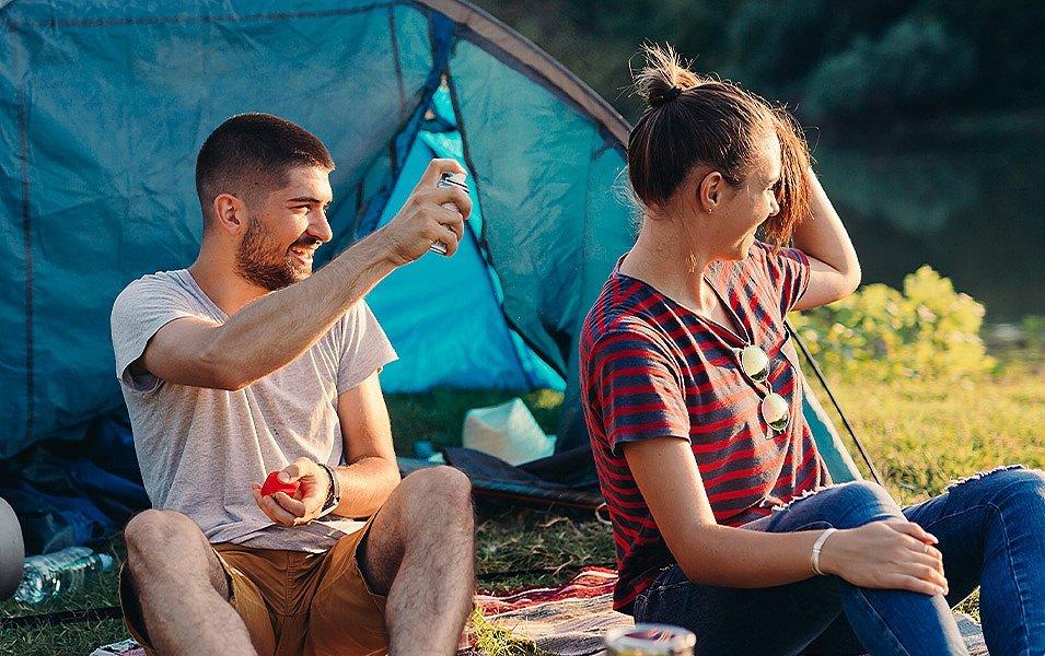 Two campers sit outside a tent in a grassy field as one sprays bug repellent on the other. 