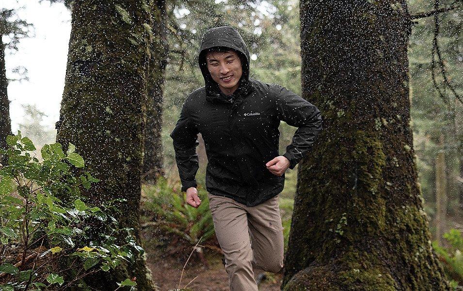 Homme courant sur un sentier forestier pluvieux portant une veste de pluie noire Columbia Sportswear.