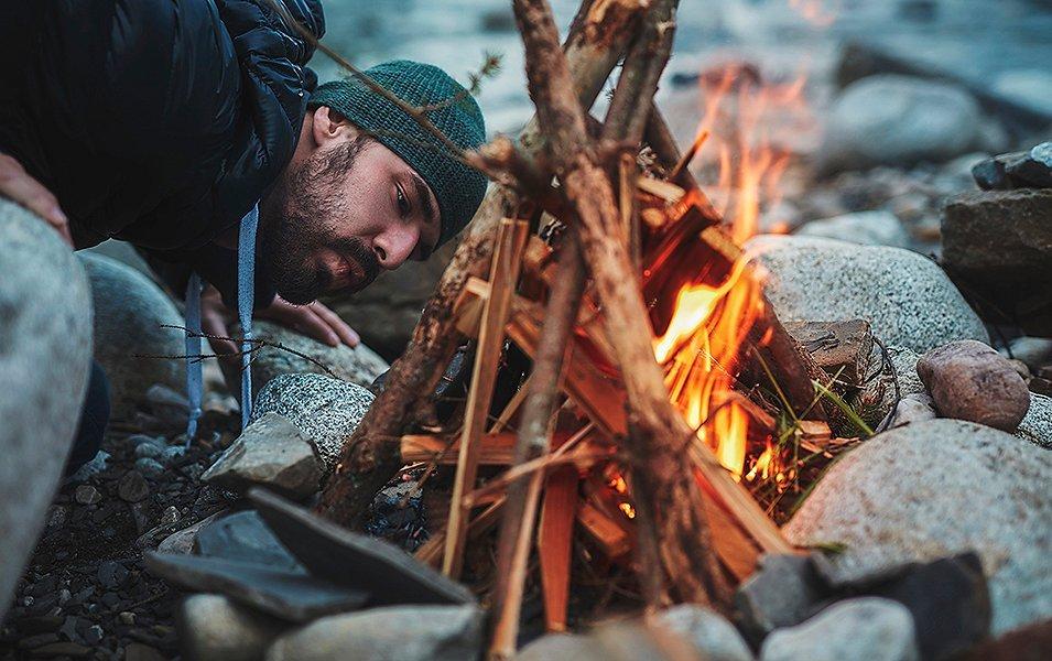 A man with a beard crouched in front of a fire blowing on it at an angle.