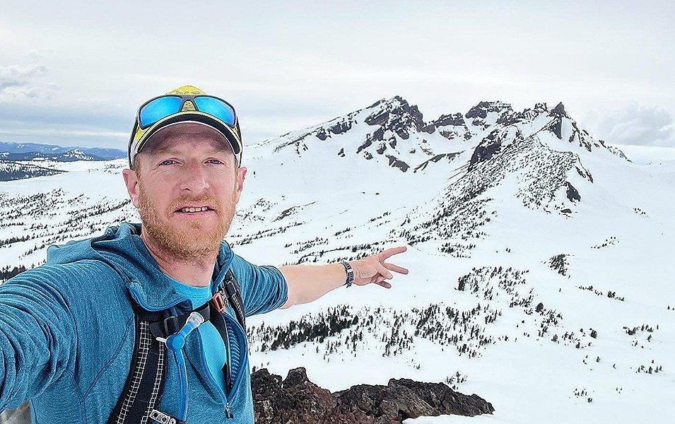 Professional trail runner Willie McBride takes a selfie on a mountain with a rugged snowy landscape in the background. 