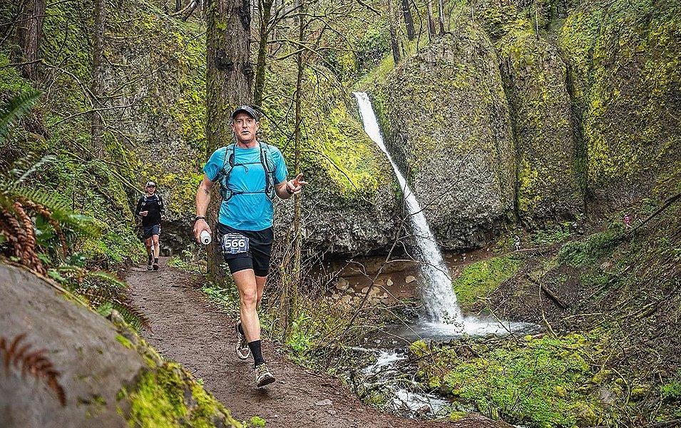 Professional trail runner Willie McBride runs through a mossy forest during an endurance race and smiles for the camera.

 