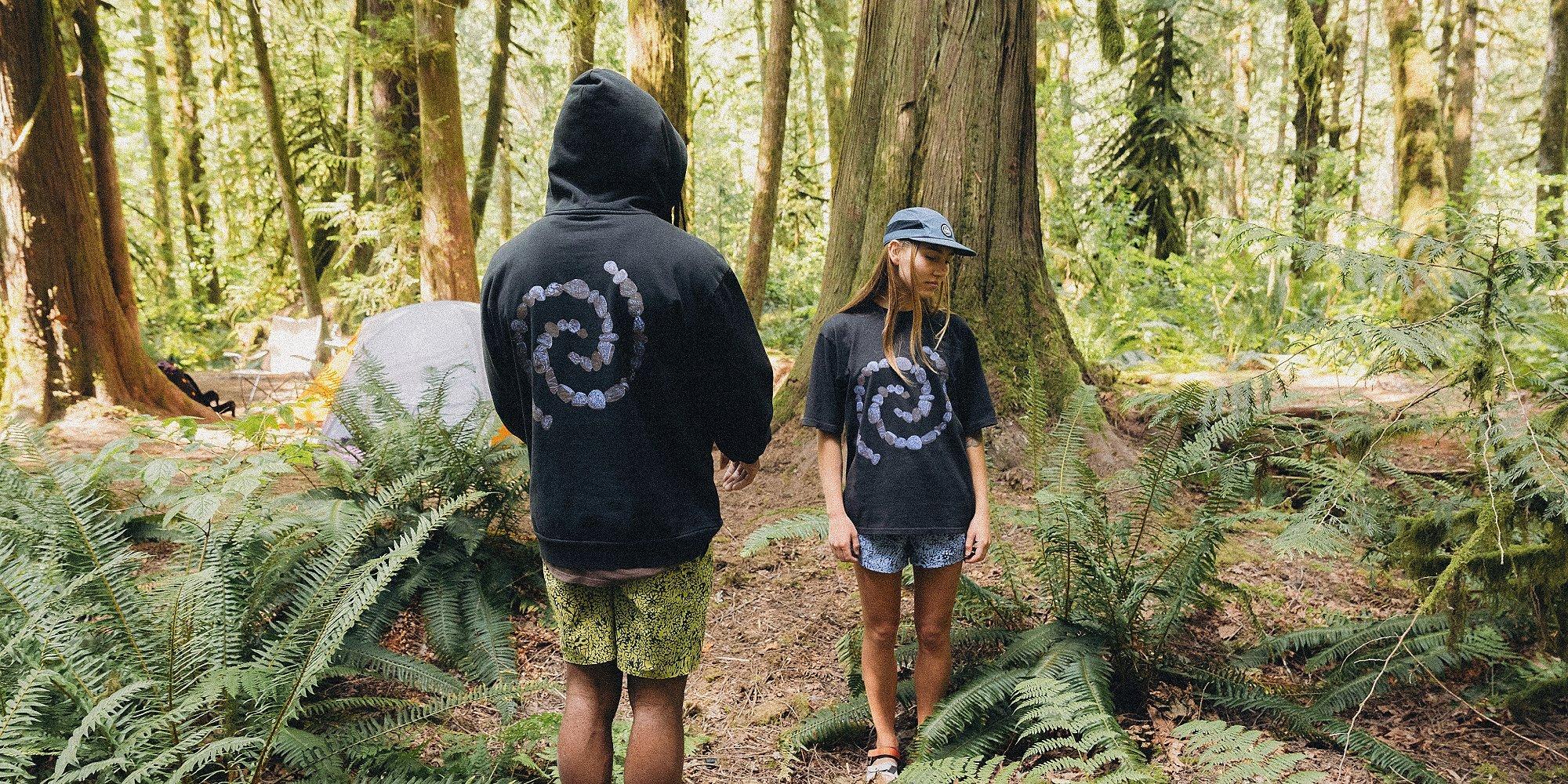 Two people wearing matching black spiral graphic shirts stand in a forest campsite surrounded by ferns and trees, with a tent and chairs in the background.