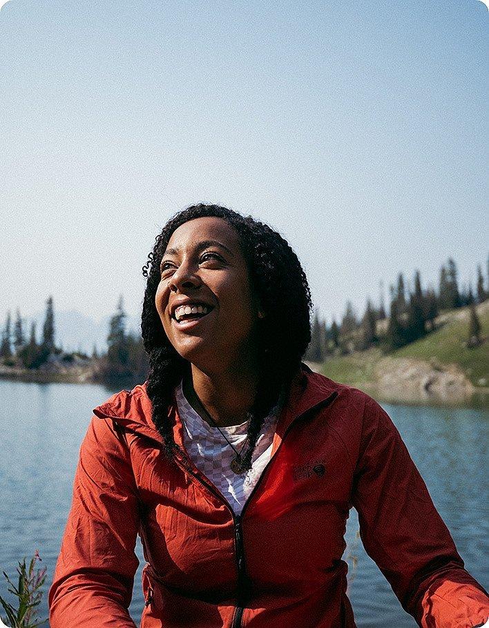 Genevive smiling looking up, in front of an alpine lake, wearing the AirShell Hoody