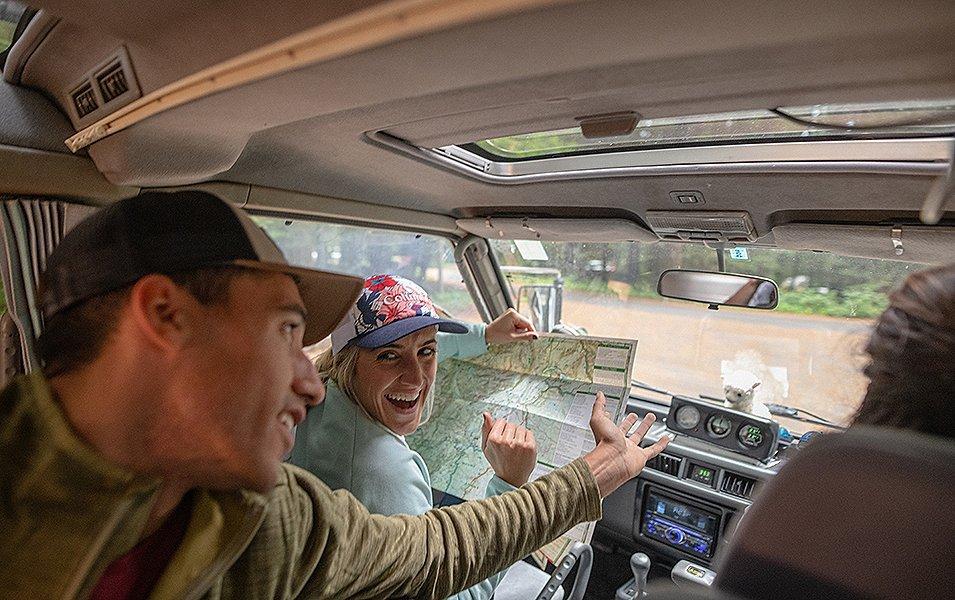 A woman in a car laughs in the driver’s seat as she reads a map while a man in the back seat reaches an arm forward. 