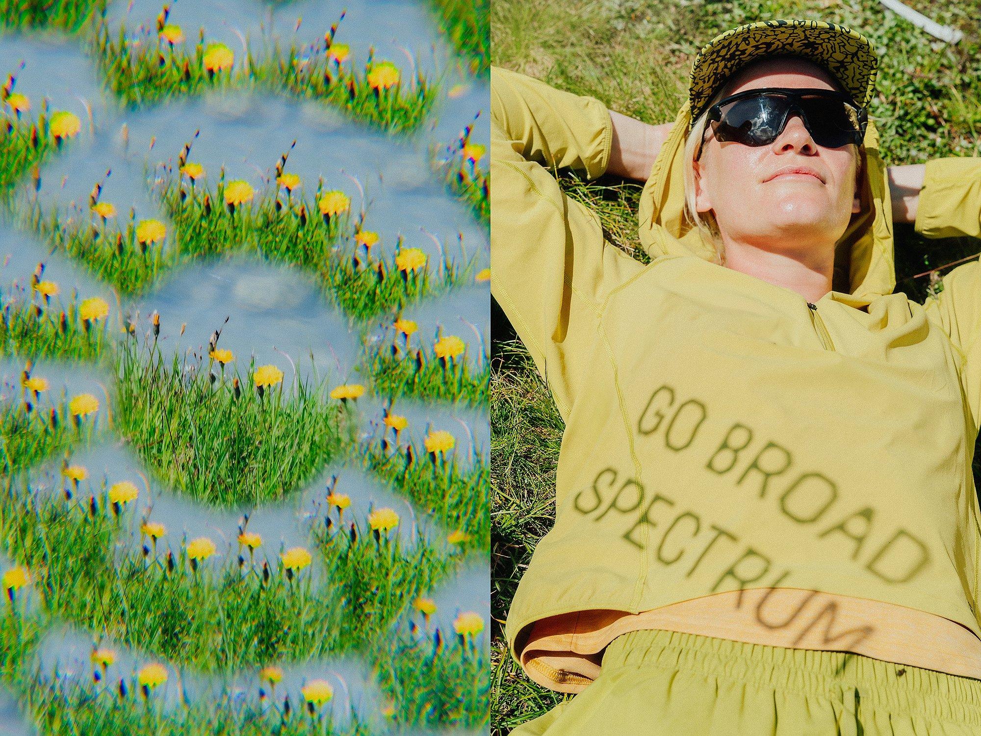A woman lounges in the grass under bright sunlight, wearing a light green Sunshield™ Hoody with the phrase "GO BROAD SPECTRUM" cast in shadow across the front. She pairs it with a matching cap and shorts, along with oversized sunglasses. Beside her, a prismatic view of a field of yellow dandelions creates a dreamy, kaleidoscopic visual effect.