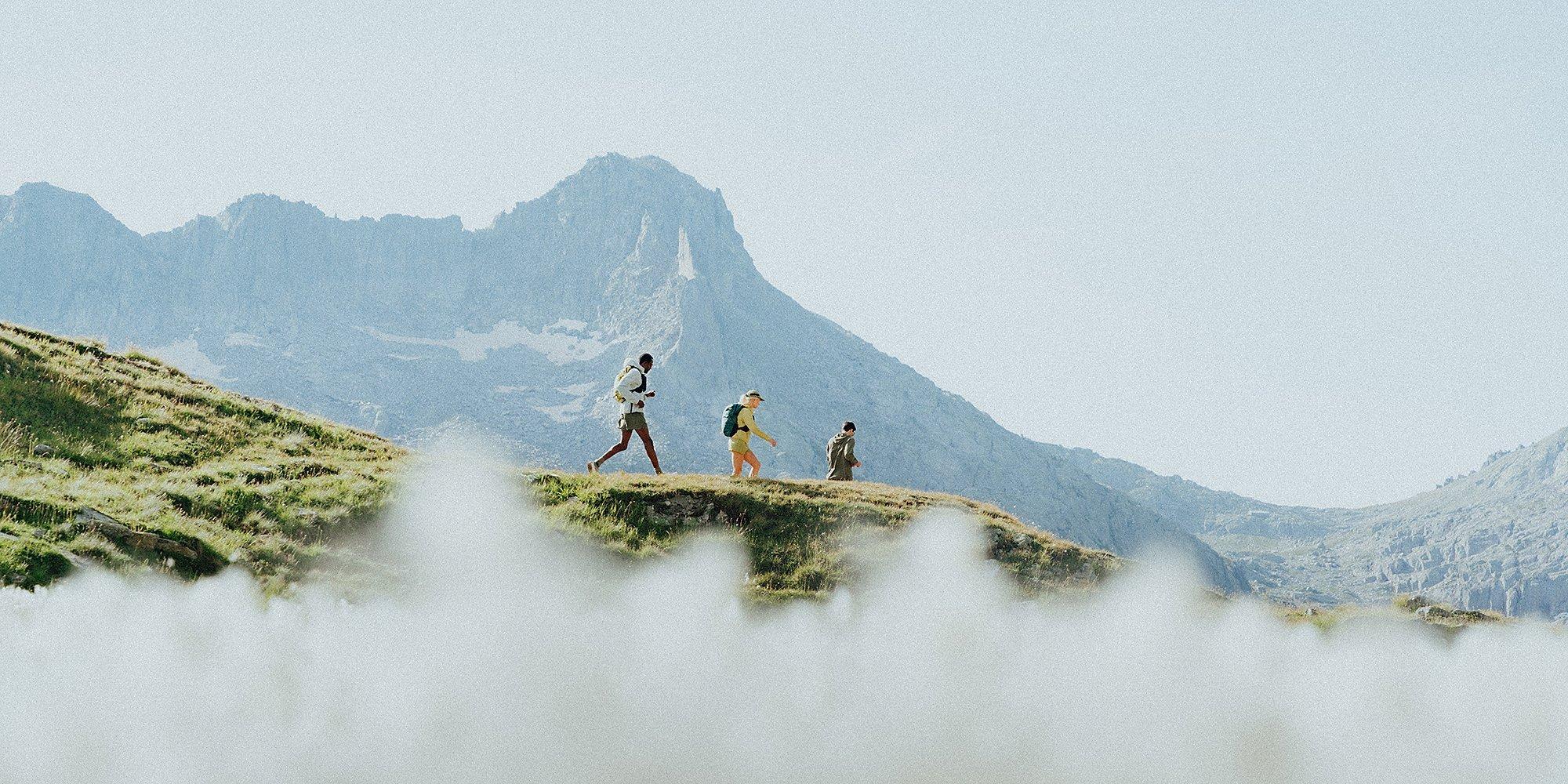 Three trail runners move along a sunlit ridgeline with dramatic mountain peaks in the background. All are wearing the Sunshield™ Hoody, ideal for lightweight protection during high-alpine adventures. Wisps of cloud or mist partially obscure the foreground, adding depth and a sense of elevation.